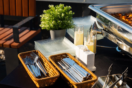 A beautifully arranged outdoor buffet includes trays with cutlery, a plant, and a warm food dish....