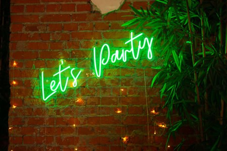 A vibrant green neon sign reads "let's party," set against a rustic brick wall adorned with string lights and lush green leaves, creating a festive atmosphere.の写真素材