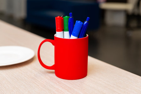 A vibrant display of assorted markers stored in a bright red mug on a wooden desk. The background shows a blurred office setting, adding a professional context.の写真素材