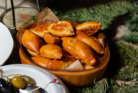 A wooden bowl lined with parchment paper is filled with assorted golden-brown baked buns and pastries, arranged on a rustic buffet table. Evergreen branches decorate the setting, and a plate with pickled vegetables and serving spoon is partially visible in the foreground.の写真素材