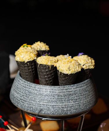 Close-up of black waffle-style cones filled with a creamy savory mixture and topped with golden crumbs, arranged upright in a gray stone bowl on a metal stand. The background is dark and softly blurred, suggesting a catered event or buffet setting.の写真素材