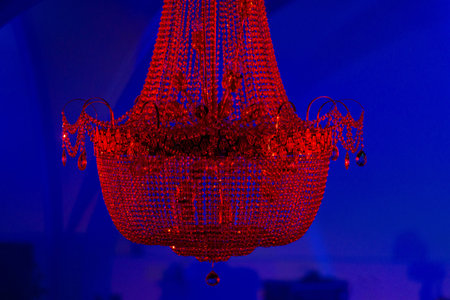 A large, ornate crystal chandelier illuminated in vivid red hangs in a dark interior, set against a deep blue background wash. The scene has a dramatic, nightclub or event-lighting atmosphere with the chandelierâs bead strands and teardrop crystals catching the light.の写真素材