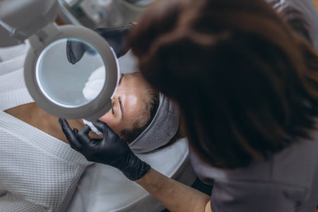 Cosmetologist in rubber gloves performs facial cleansing using magnifying glass with LED light lupa. High quality photoの写真素材