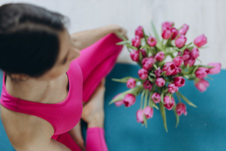 Top view of a woman in a pink sports suit sitting cross-legged on a yoga mat, with a bouquet of pink tulips beside her. High quality photoの写真素材