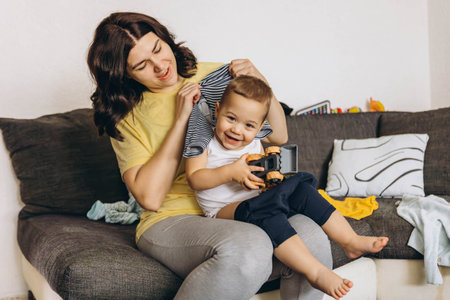 Mother changing her baby, while the baby laughs, holding a toy car in his hand. High quality photo