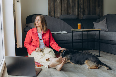 Freelancer woman working from home with laptop, sitting on floor in cozy living room, barefoot and relaxed, petting her dog. Remote work and pet-friendly lifestyle concept. High quality photoの写真素材