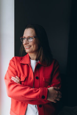 Business portrait of a confident woman in red jacket and glasses standing by the window, arms crossed, smiling. Concept: leadership, personal brand, online expert, business style. High quality photoの写真素材