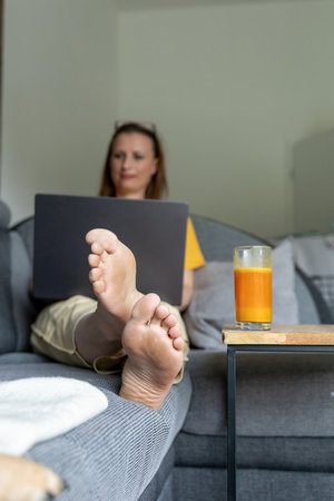 Smiling woman working remotely on laptop while relaxing on the couch at home. Freelance lifestyle, comfort, work-from-home routine, and modern digital communication. High quality photoの写真素材
