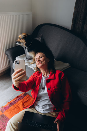 Woman working remotely with laptop, taking a selfie with her dog at home. Happy freelancer enjoying break with pet, sitting on floor in cozy living space, casual digital lifestyle. High quality photoの写真素材