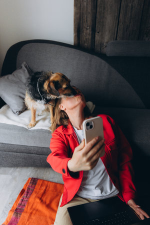 Woman working remotely with laptop, taking a selfie with her dog at home. Happy freelancer enjoying break with pet, sitting on floor in cozy living space, casual digital lifestyle. High quality photoの写真素材