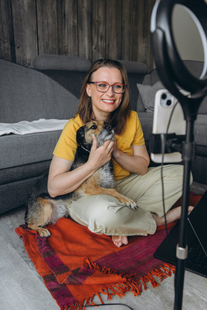 Smiling woman with glasses holding her dog while recording a video call at home. Cozy interior,...