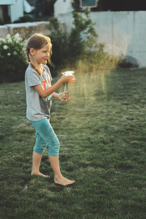 Smiling little girl playing with water spray bottle on green grass in backyard. Summer fun, wet clothes, carefree childhood moment outdoors in the evening lightの写真素材