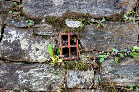 Ancient wall made of natural rocks with window-like water-drain pipe in the center and with plants and lichens between blocks as a backgroundの写真素材