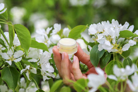 Close up of the moisturizing skincare cream in an open small glass jar in woman's hand among the...