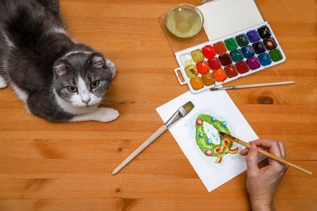 An unrecognizable woman painting Christmas garland at her home with her cat watching her work. Photo from aboveの写真素材