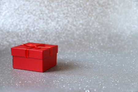 Red gift box with red and gold ribbon on silver sparkling glittery background. Photo in perspective...