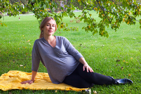 Pregnant woman sitting in the park on the yellow knitted plaid in warm summer or autumn day