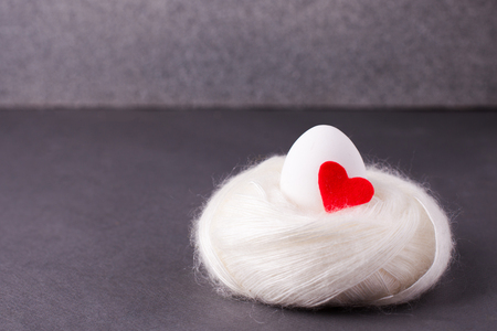 Easter concept, the origin of life, love and purity - a white egg with a red heart lies in a soft cozy warm nest on a gray backgroundの写真素材