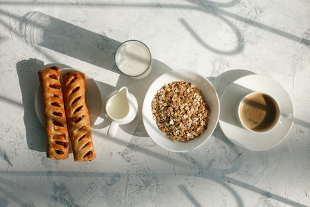 Breakfast from oatmeal with milk and cup of black coffee and rolls with jam and sunlight ray shadow on a gray table. Free space for text . Top viewの写真素材
