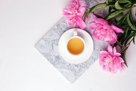 Fashion blogger styled desk frame with flowers, cup of herbal tea, pink peonies on marble plate on white, breakfast morning lifestyle, minimal concept, copy spaceの写真素材