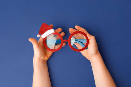 glasses Santa's Christmas in kid hands on classic blue background. Masquerade festive party new year funny sunglasses, trendy minimal winter holiday idea, banner, flyer coupon, party invitationの写真素材
