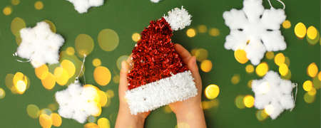 Kids hands holding decorative red Christmas hat on natural green background. Sale, Holiday, greering...