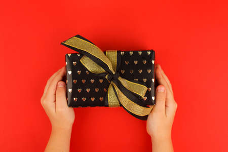 Kids hands holding decorativegift box with bow on red background. Sale, Holiday, greering card Happy new year 2022, Merry Christmas, banner for your site, invitation, flyer, holiday invitationの写真素材