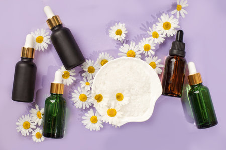 Dark glass cosmetic bottles with water drops on pastel purple violet background with chamomile flowers. Concept of natural moisturizing cosmetics for skin care, organic bio skincare flyerの写真素材