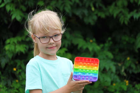 a blonde little girl playing with an anti-stress multi-colored pop toy on the street, child against...