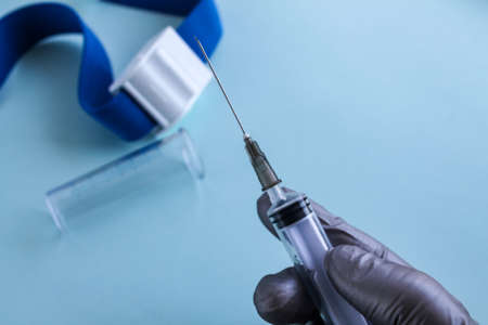 A hand in a gray medical glove holds a syringe before taking blood from a vein, next to it on a blue background lie a test tube and a medical venous tourniquetの写真素材