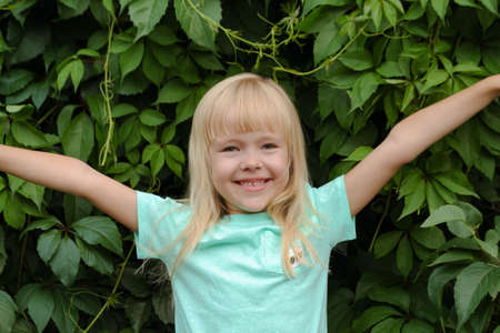 blonde little girl smiles, shows joy, emotions of the child against the background of green leaves.の写真素材