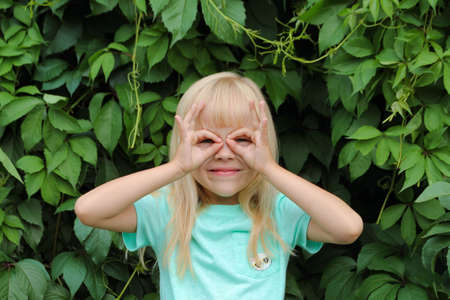 A little blonde girl makes glasses or a  mask on her face with her hands. The child makes faces against the background of green leaves.の写真素材