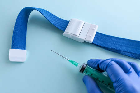 A hand in a blue medical glove holds a syringe before taking blood from a vein, a medical venous tourniquet lies next to it on a light blue backgroundの写真素材