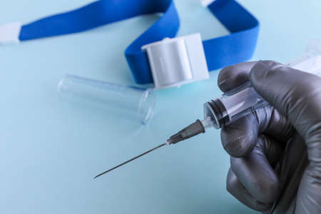 A hand in a gray medical glove holds a syringe, a test tube and a tourniquet for taking blood from a vein are lying next to it. light blue background, close-up.の写真素材