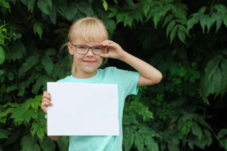 Blonde little girl with glasses holding a blank sheet of paper with space for text, the child against the background of green leaves.の写真素材