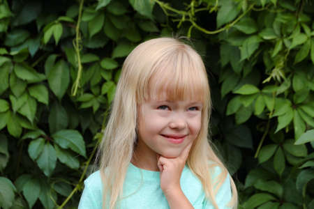 a little blonde girl in a mint T-shirt thinks about something joyful or cheerful, happily dreams. Brooding child against the background of green leaves.の写真素材