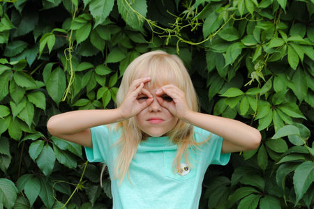 A little blonde girl makes glasses or a mask on her face with her hands. The child makes faces against the background of green leaves.の写真素材