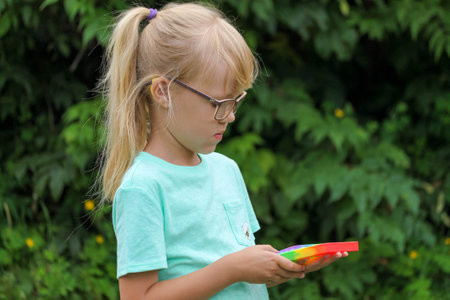 a blonde little girl playing with an anti-stress multi-colored pop toy on the street, child against the background of green leaves.の写真素材
