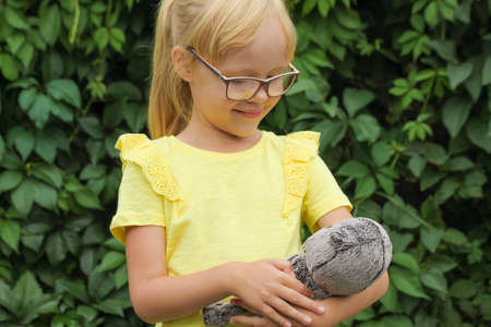 Blonde girl in glasses and a yellow T-shirt with a favorite toy kitten baby Basik in the garden against the background of greenery.の写真素材