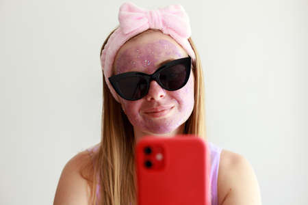 woman in a cosmetic mask on her face and glasses with a red smartphone on a white backgroundの写真素材