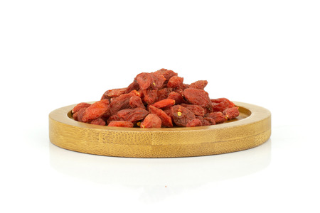 Lot of whole dried red goji berries on round bamboo plate isolated on white background