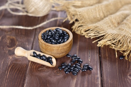 Lot of whole raw black turtle beans jute cloth and a scoop with wooden bowl on brown woodの写真素材