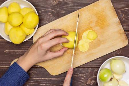 Woman hands are cutting potatoes. recipe step by step baked potatoes with onion on chopping board flatlay on brown woodの写真素材