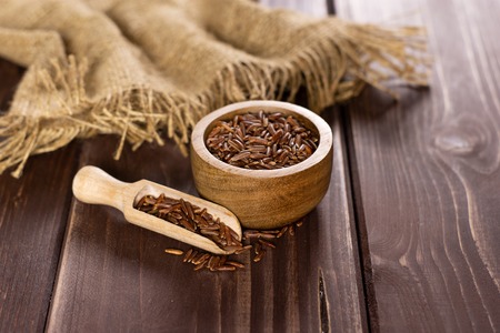 Lot of whole raw red rice jute cloth and a scoop with wooden bowl on brown woodの写真素材