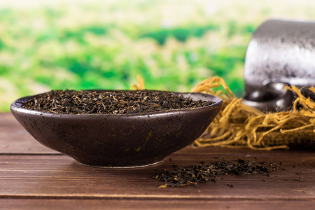 Lot of pieces of dry black tea earl grey in a grey ceramic bowl on jute cloth with black ceramic cup green tea field behindの写真素材