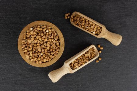 Lot of whole hulled raw brown buckwheat grain in a wooden bowl with wooden scoop flatlay on grey stoneの写真素材