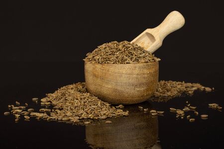 Lot of whole dry caraway fruits in a wooden bowl with wooden scoop isolated on black glass