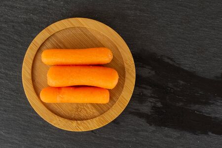 Group of three whole peeled orange baby cut carrot on bamboo plate flatlay on grey stoneの写真素材