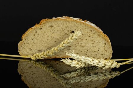 One half of fresh baked rye wheat bread with three golden wheat ears isolated on black glass