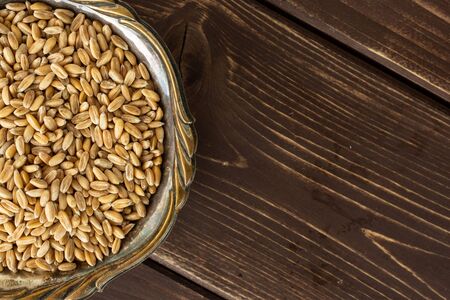 Lot of whole fresh beige dinkel wheat grain in old iron bowl flatlay on brown wood